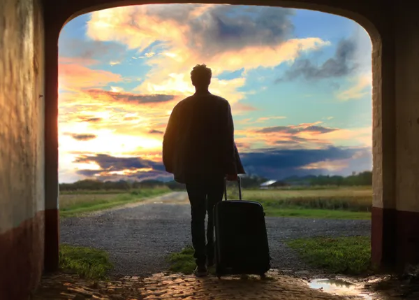 A silhouette of a man holding a suitcase walking through an arched entryway towards sunrise.