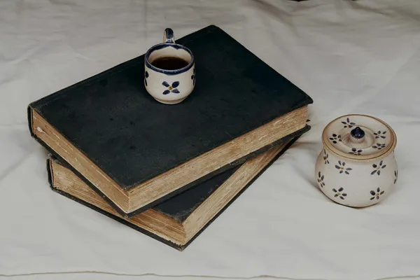 A small cup on top of two, weathered, blue books, with a small teapot besides it